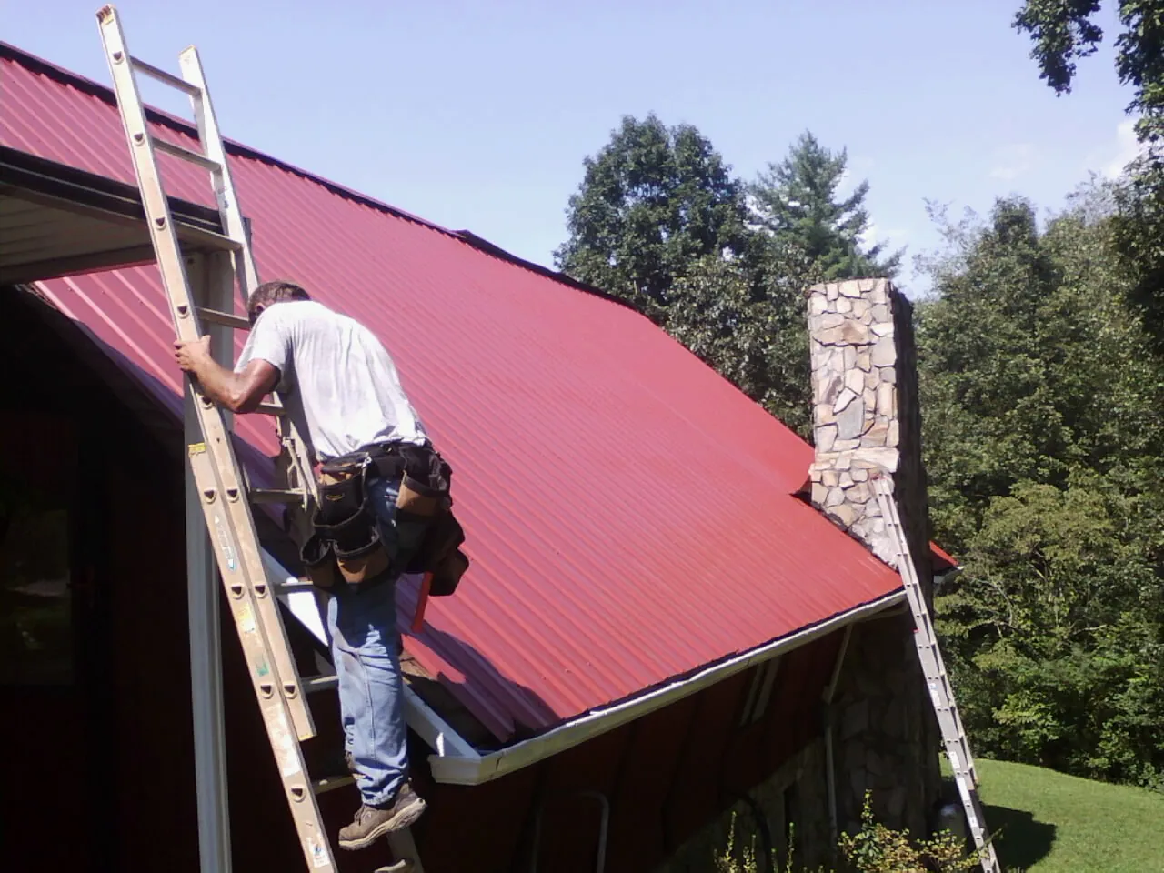 Roofer on a ladder beside a red metal roof and stone chimney — Soffit & Fascia Repair by Roofing Brothers in Mc Kenzie