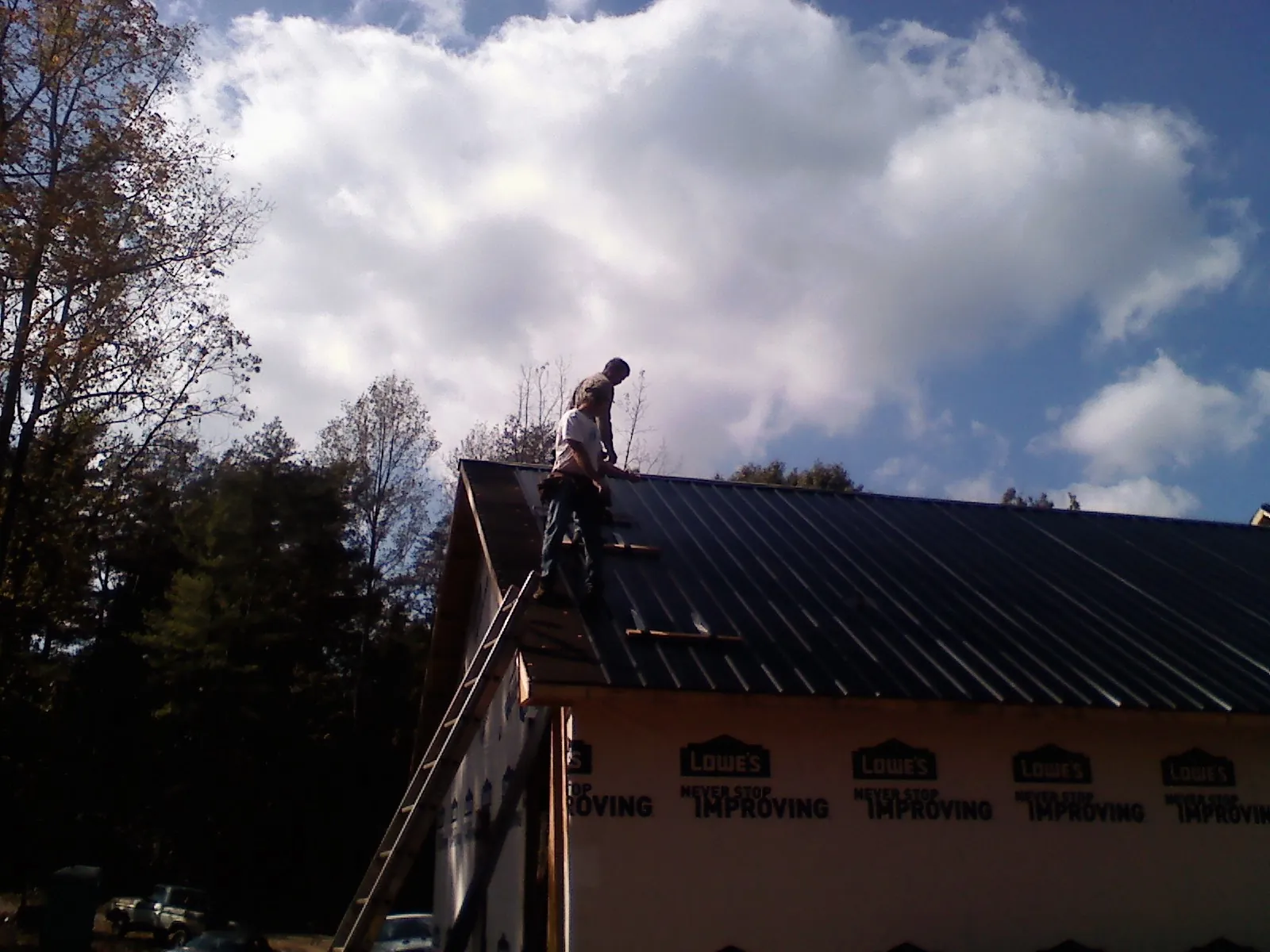 Roofer fastening dark metal panels high against a cloudy sky, from a Roof Inspection job in Mc Kenzie